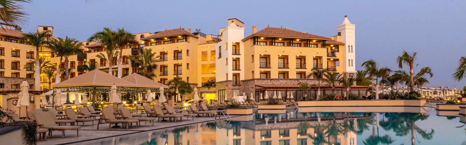Hotel-Schwimmbad bei Sonnenuntergang auf Teneriffa, mit Liegeplätzen und Palmen rundherum.