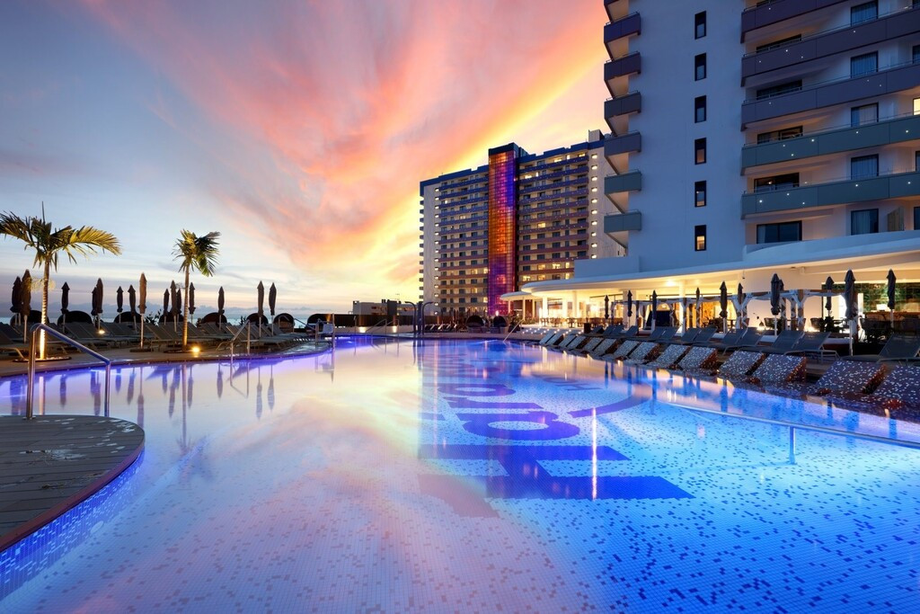Schwimmbad, das bei Sonnenuntergang in einem Hotel auf Teneriffa erleuchtet ist, umgeben von Sonnenliegen und Palmen.