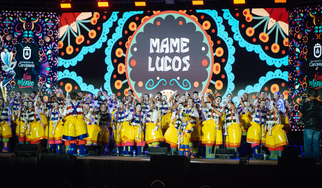 Gruppe von Menschen auf der Bühne von Candelaria, mit farbenfrohem Hintergrund und Text Mame Luicos.