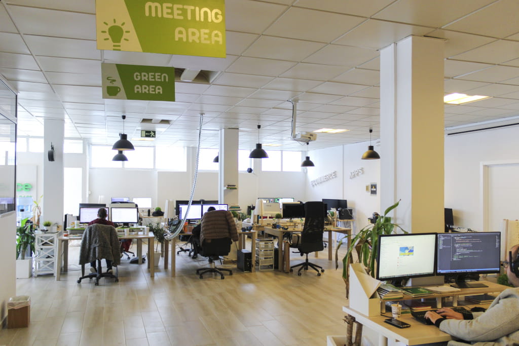 Großraumbüro mit Computern und Mitarbeiter, Schilder MEETING AREA und GREEN AREA.