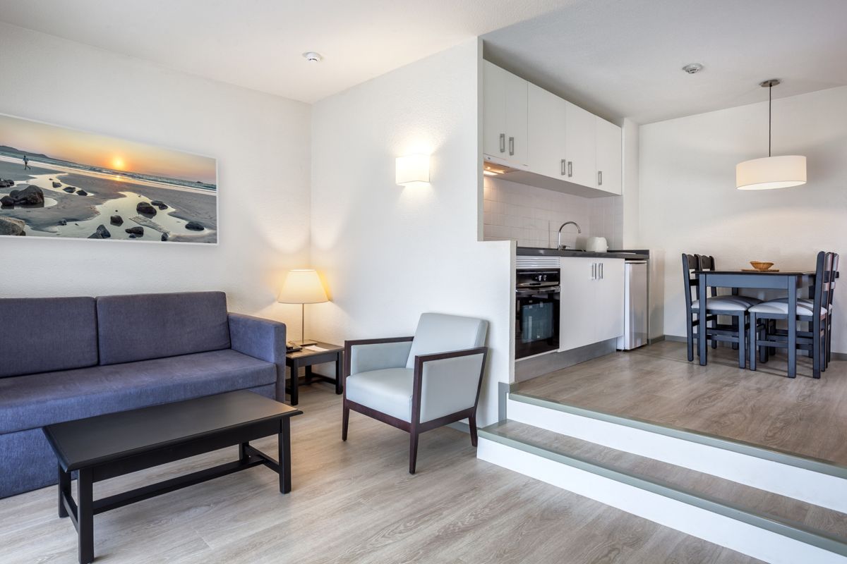 Wohnzimmer mit Sofa, Sessel, Tischlampe und Küche in einer Ferienwohnung in Teneriffa.
