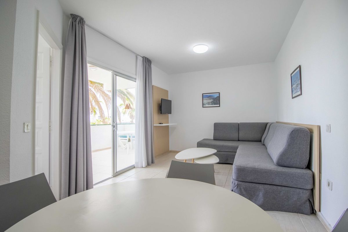 Wohnzimmer in Teneriffa mit grauem Sofa, Tisch, Gemälden und Balkonzugang.