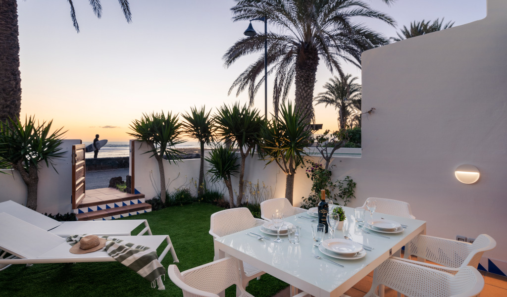 Terrasse mit Tisch und Stühlen, Palmen, Surfer am Tor, Strand von Tenerife im Hintergrund.