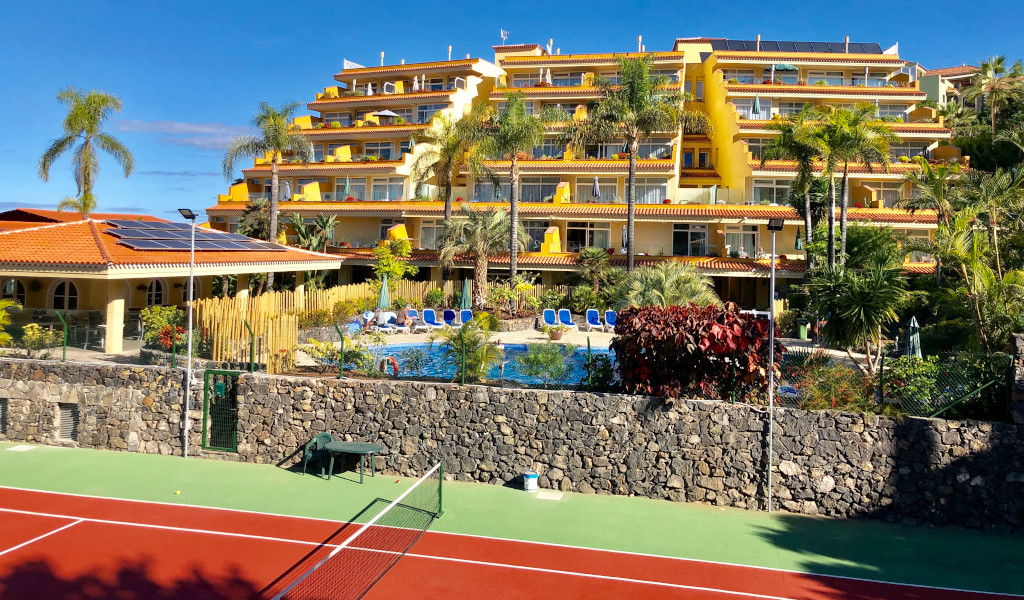 Gebäude mit Pool und Palmen in Puerto de la Cruz, Teneriffa, mit Tennisplatz im Vordergrund.