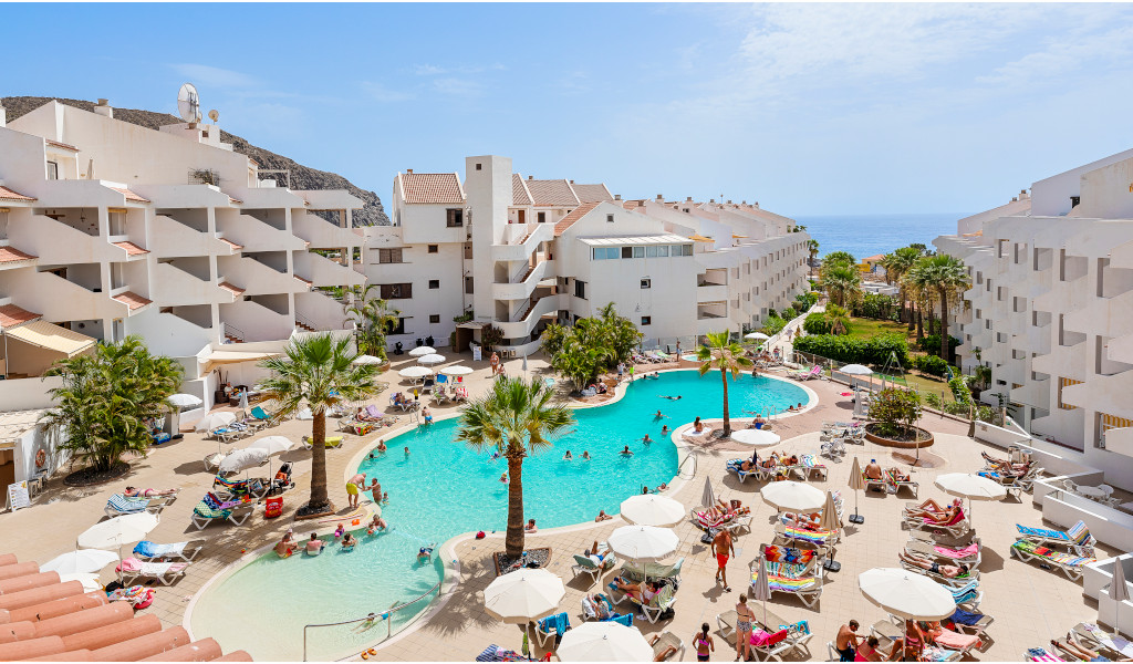Poollandschaft und Apartments in Los Cristianos, Teneriffa, mit Meerblick im Hintergrund.