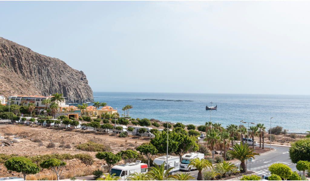 Küste von Los Cristianos mit Meerblick und Schiff, umgeben von Palmen und Gebäuden.