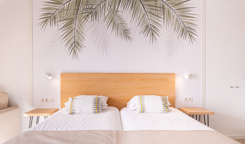 Zwei Betten mit hellem Holzkopfteil und grauen Palmenblättern an der Wand in Teneriffa.