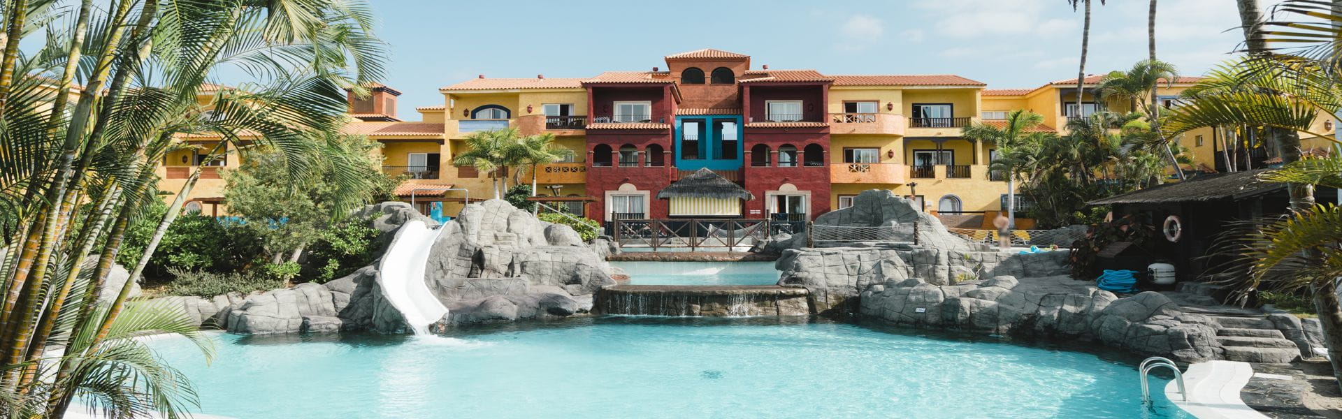 Hotel mit Rutschen und Pool in Teneriffa, umgeben von Palmen und Gebäuden.