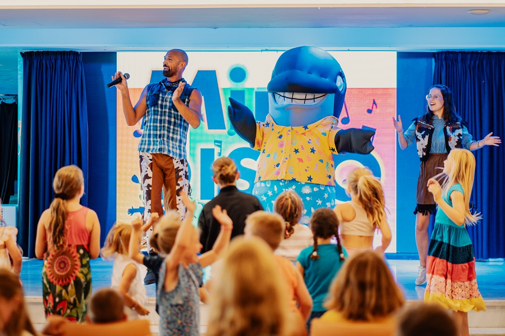 Kinder und Animateure tanzen und singen mit einem Wal-Maskottchen auf einer Bühne in Teneriffa.