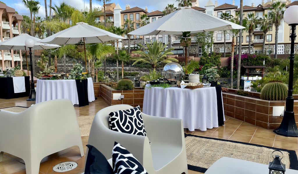 Terrasse in Adeje mit Tischen, Stühlen und Sonnenschirmen, umgeben von Palmen.