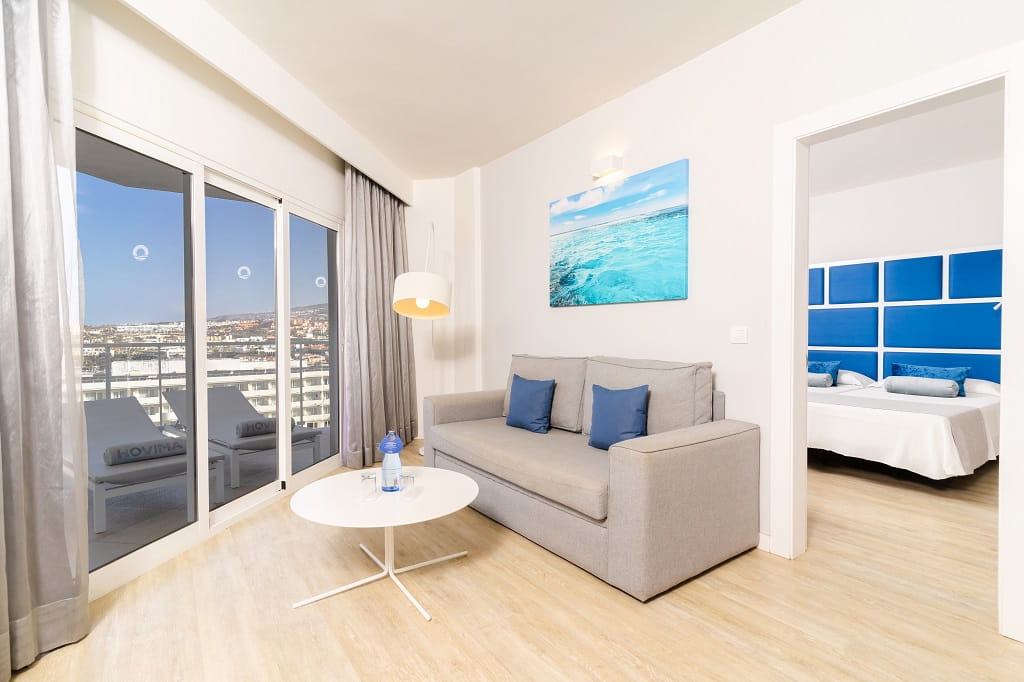 Wohnbereich mit grauem Sofa, Blick auf Balkon und Schlafzimmer im Hotelzimmer in Teneriffa.