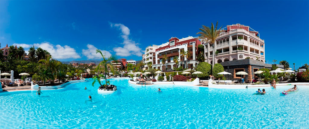 Großes Hotel mit Pool und Palmen in Costa Adeje, Teneriffa, unter blauem Himmel.