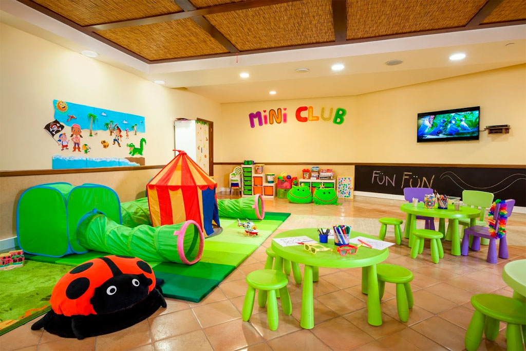 Spielzimmer im Mini Club Tenerife mit Spielzeug, Tischen und Fernseher.