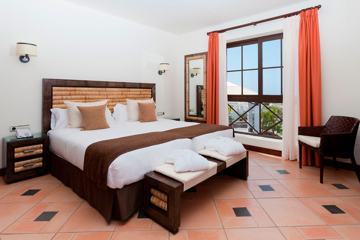 Schlafzimmer mit Doppelbett, Fensterblick auf Teneriffa, orange Vorhänge und helle Wände.