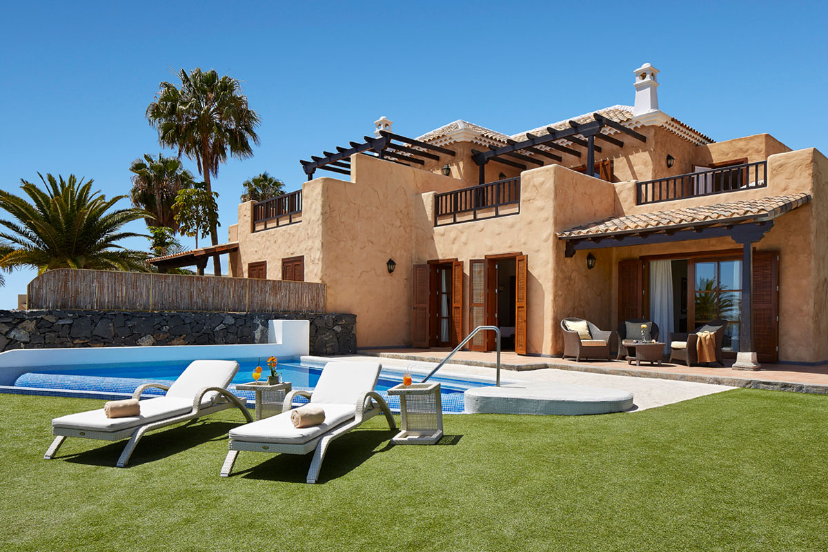 Zwei Liegestühle am Pool vor einer Villa in Teneriffa mit Palmen und blauem Himmel.