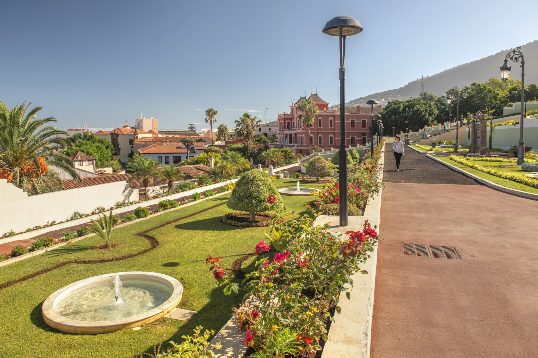 Gärten und rotes Gebäude in Puerto de la Cruz, Teneriffa, mit Bergen im Hintergrund.