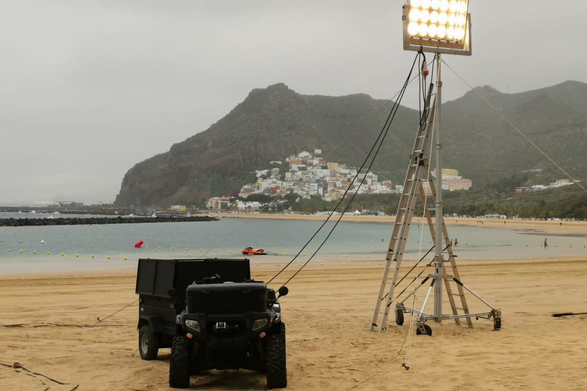 Lichter und Fahrzeuge am Strand von Las Teresitas, Teneriffa, mit Häusern und Bergen im Hintergrund.