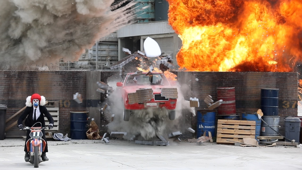 Motorradfahrer mit Clownshelm und rotem Auto bei Heckexplosion.
