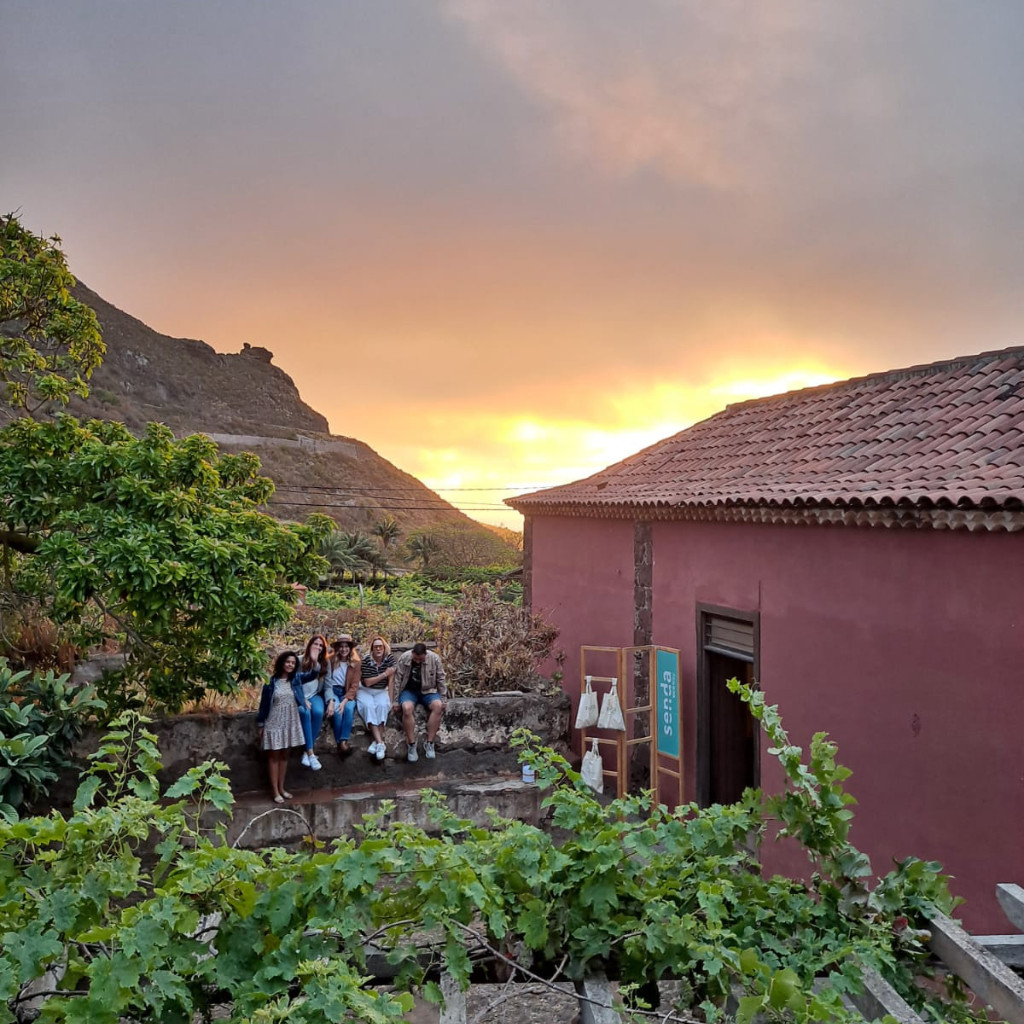 Eine Gruppe von Menschen sitzt bei Sonnenuntergang in einem Garten auf Teneriffa; ein Schild weist einen Weg.