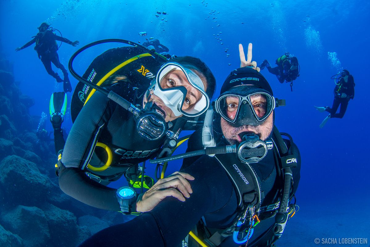 Taucher, die unter Wasser auf Teneriffa lächeln, mit anderen im Hintergrund.