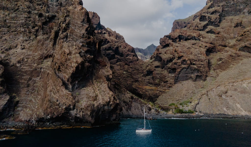 Segelboot an der felsigen Küste von Teneriffa mit blauem Wasser und Bergen im Hintergrund.