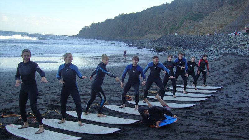 Eine Gruppe von Menschen lernt das Surfen am Strand auf Teneriffa, aufgereiht auf Brettern.