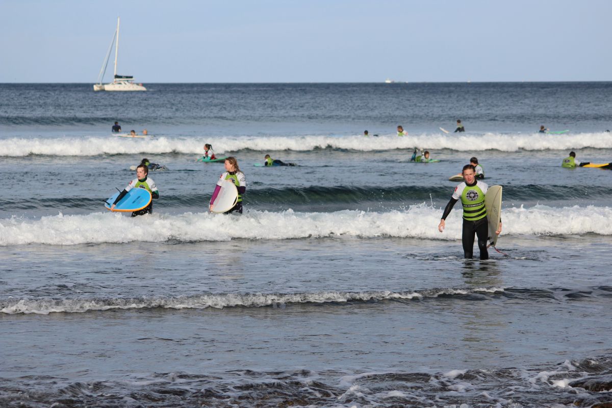 Surfer mit Brettern in Playa de Las Américas, Teneriffa, mit einem Segelboot im Hintergrund.