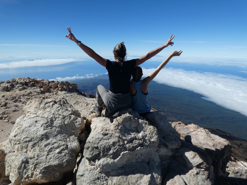 Menschen heben ihre Arme auf dem Gipfel des Montes Teide auf Teneriffa und blicken auf die wolkige Landschaft.