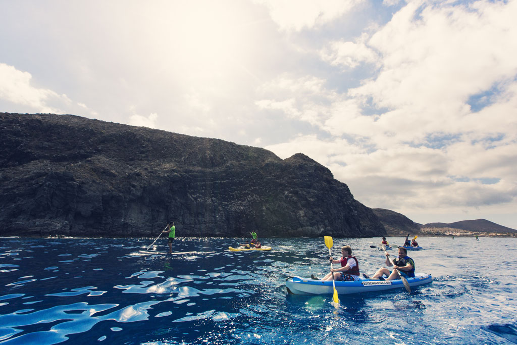 Menschen, die an der Küste Teneriffas Kajak und Paddle-Surfen mit Bergen im Hintergrund unterwegs sind.