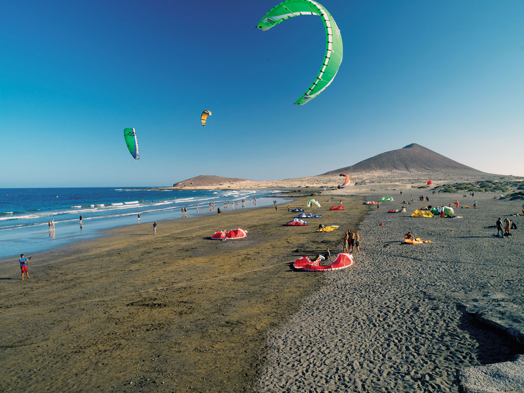 Menschen kitesurfen am Strand von El Médano auf Teneriffa, mit Bergen im Hintergrund.