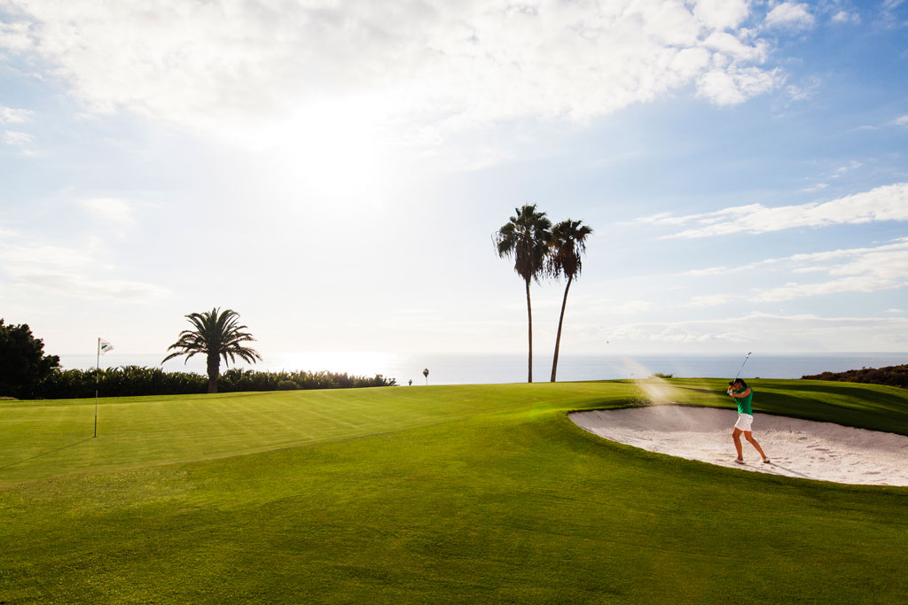 Person, die auf einem Golfplatz an der Küste Teneriffas mit Palmen und klarem Himmel Golf spielt.