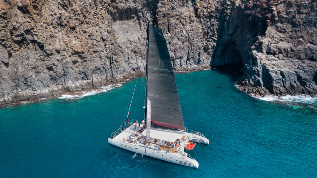 Katamaran, der in der Nähe von felsigen Klippen an der Küste Teneriffas segelt.