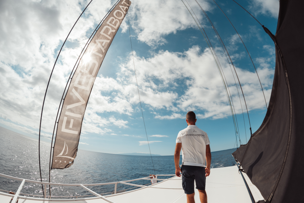 Person am Bug eines Bootes trägt ein T-Shirt mit der Aufschrift FIVESTARBOAT, blickt aufs Meer auf Teneriffa.
