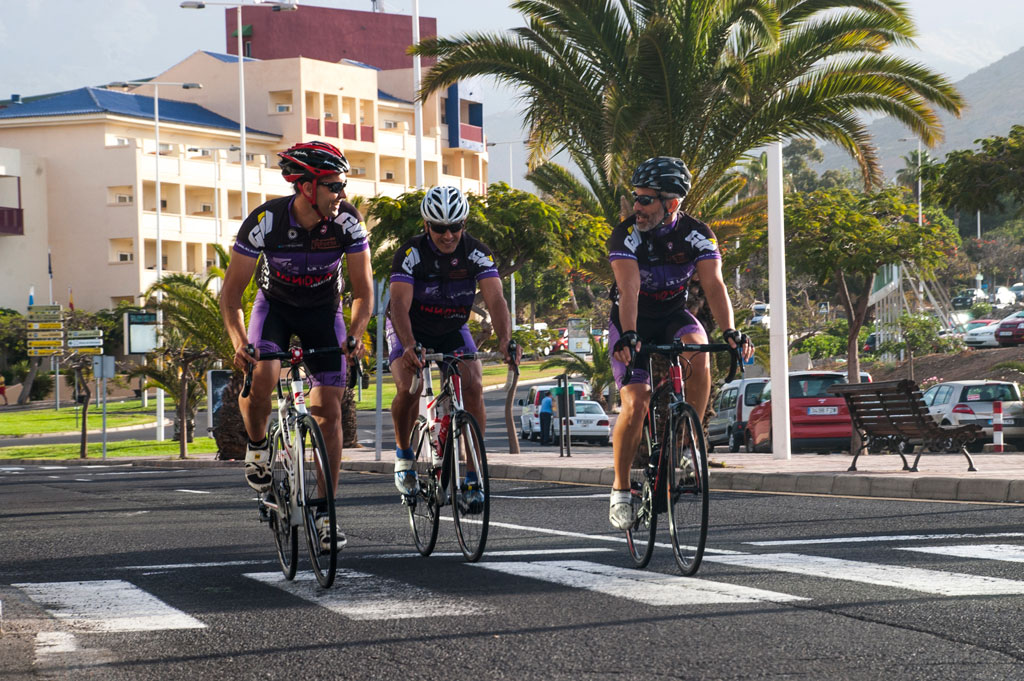 Radfahrer überqueren eine Straße auf Teneriffa mit einem Gebäude im Hintergrund und Palmen drumherum.