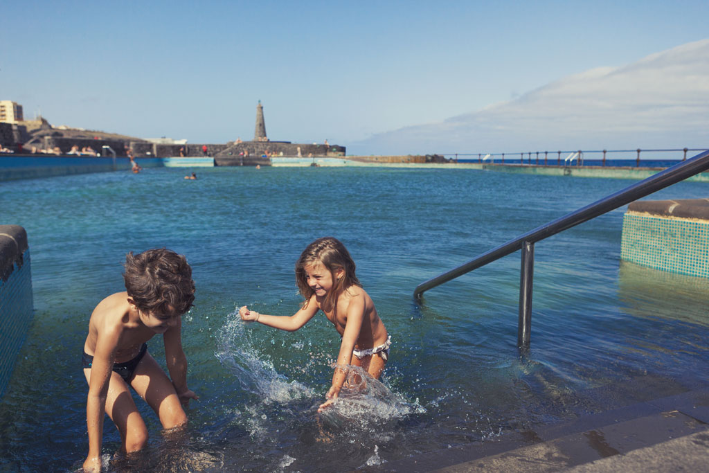 Kinder, die in einem Naturbecken in Bajamar, Teneriffa, spielen, mit einem Leuchtturm im Hintergrund.