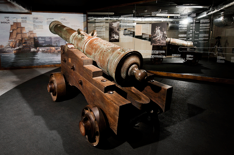 Antike Kanone ist in einem Museum auf Teneriffa ausgestellt, mit Informationstafeln dahinter.