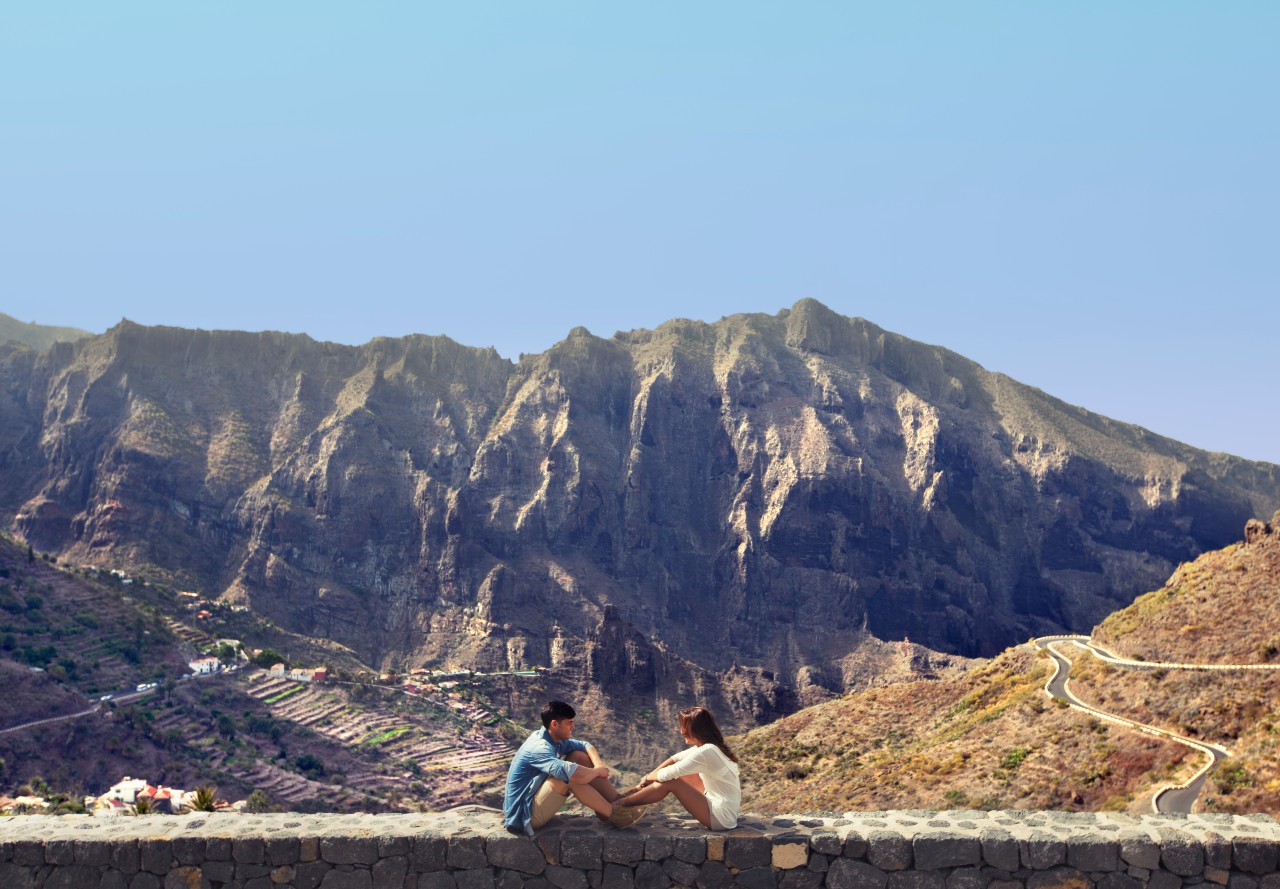 Menschen, die vor den Masca-Bergen auf Teneriffa sitzen, mit einem gewundenen Pfad im Hintergrund.