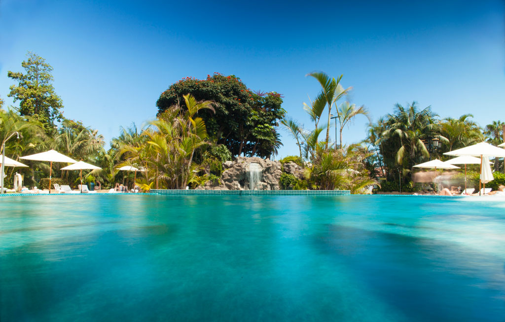 Schwimmbad mit Sonnenschirmen und tropischer Vegetation in einem Hotel auf Teneriffa.